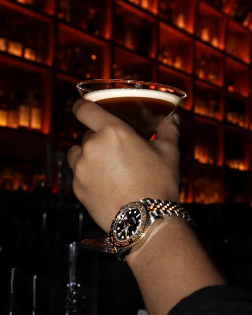 An espresso martini raised in front of the backlit bottle wall